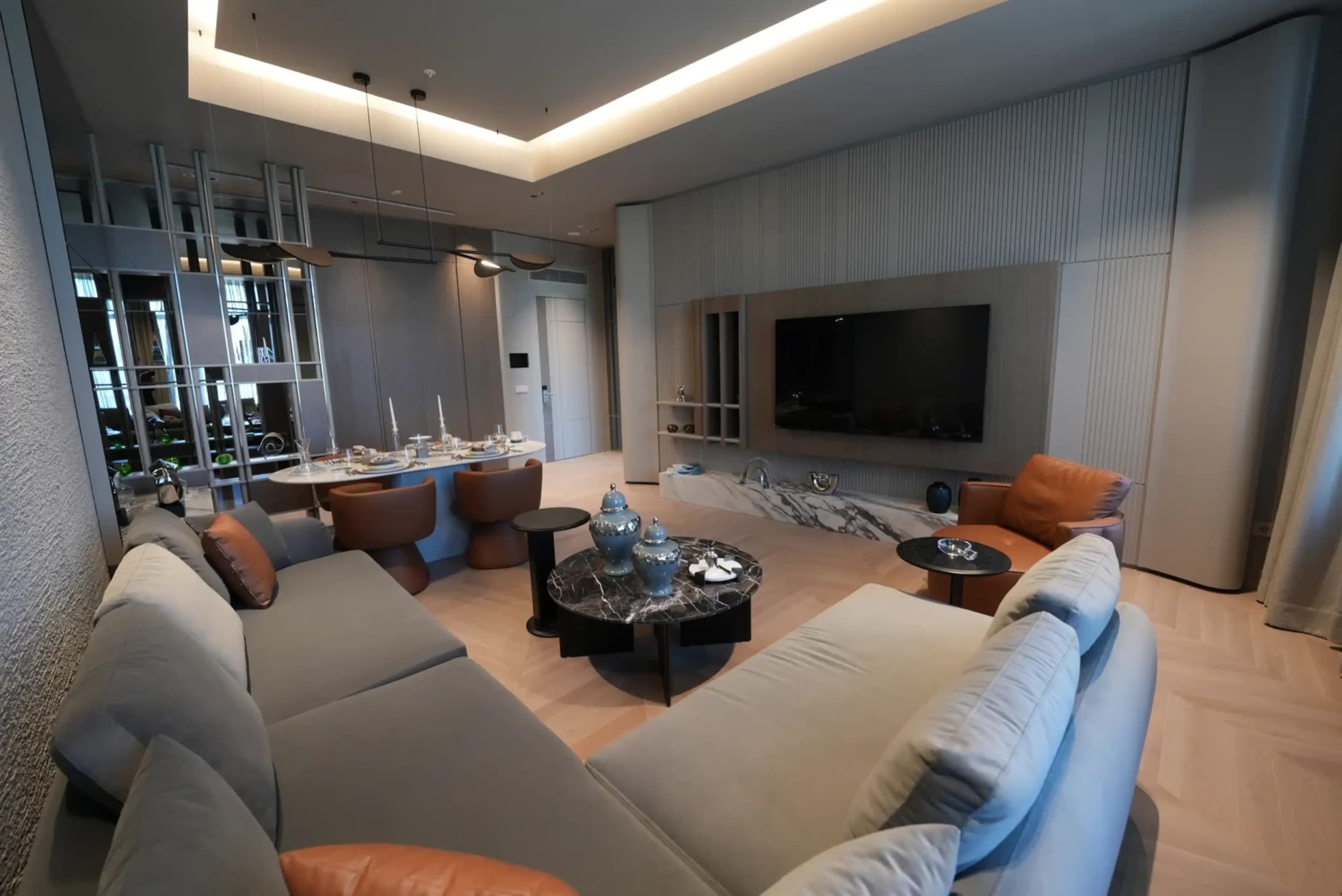 Living room of a furnished residence with a wall-mounted television, dining table beyond and chevron parquet flooring