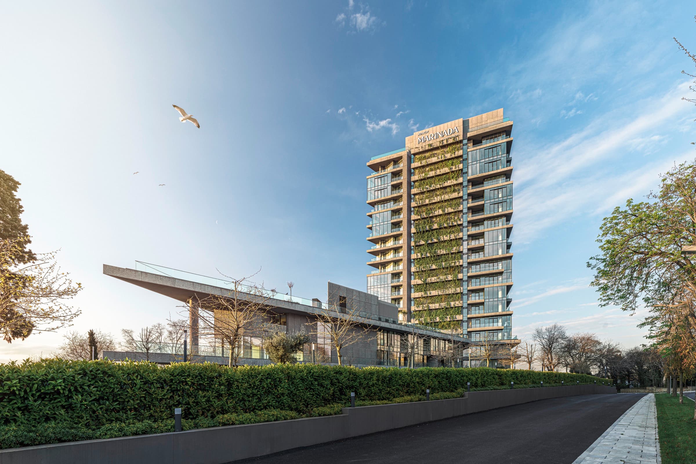 Ataköy Marinada Residence'a yaklaşım: budanmış çitin yanından geçen taş döşeli yol alçak konsol giriş pavyonuna varıyor, arkada dikey bahçesiyle kule yükseliyor ve gökyüzünü bir martı geçiyor