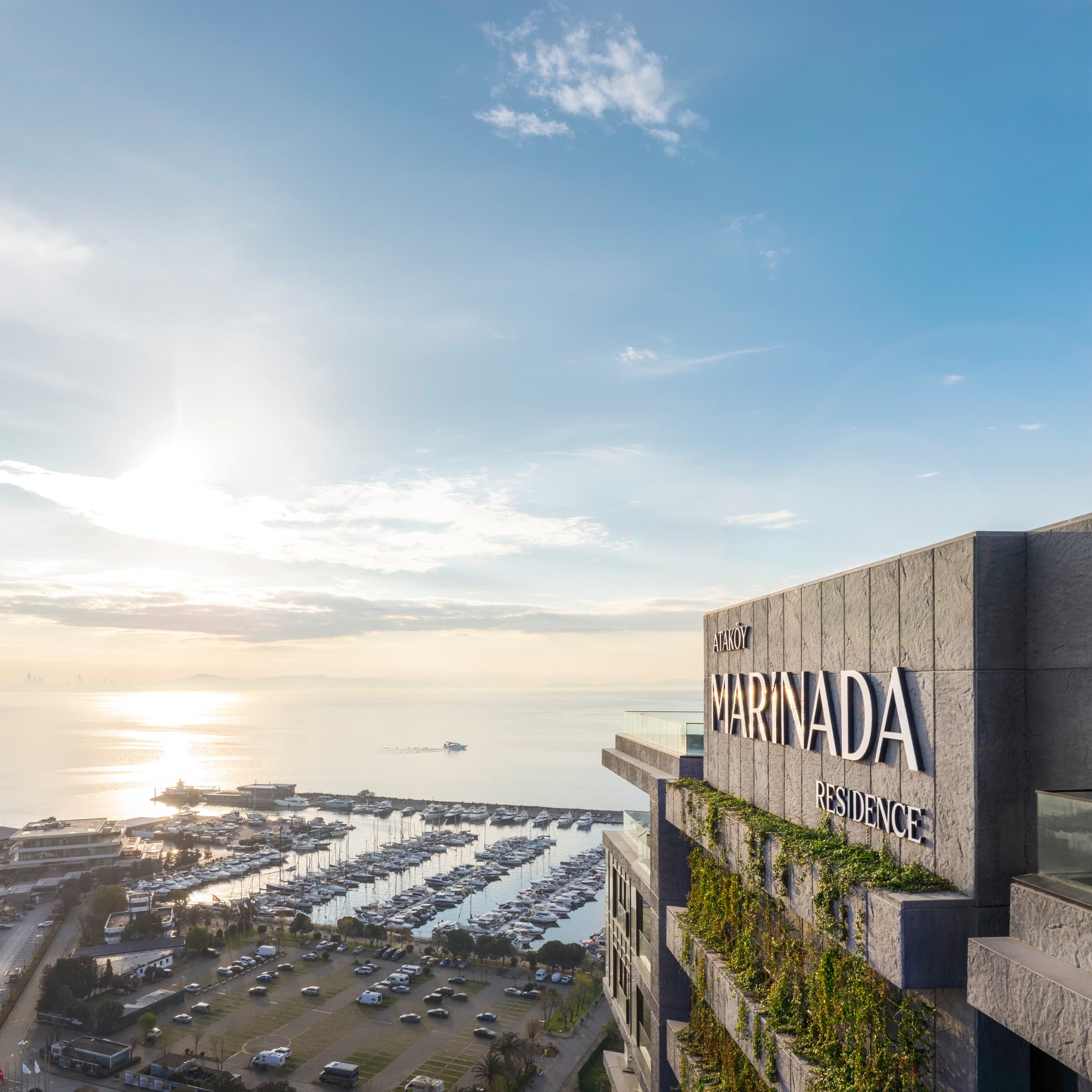 Ataköy Marinada Residence'ın gün doğumundaki korkuluğu: taş yüzeyinde bitkilendirilmiş terasların üzerinde ATAKÖY MARINADA RESIDENCE harfleri, aşağıda marina bağlama yerleri, otopark ve Marmara Denizi'nin üzerinden doğan güneş