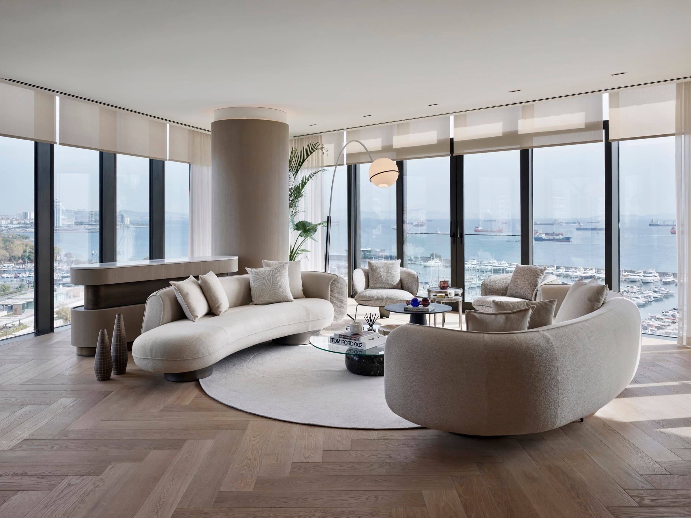 Ataköy Marinada Residence'ta bir rezidansın salonu: balık sırtı meşe parkenin üzerindeki yuvarlak halıda krem kavisli kanepeler ve bukle berjerler, ortada yuvarlatılmış bir kolon, iki cephede tam boy cam marinaya ve Marmara Denizi'ndeki gemi trafiğine bakıyor