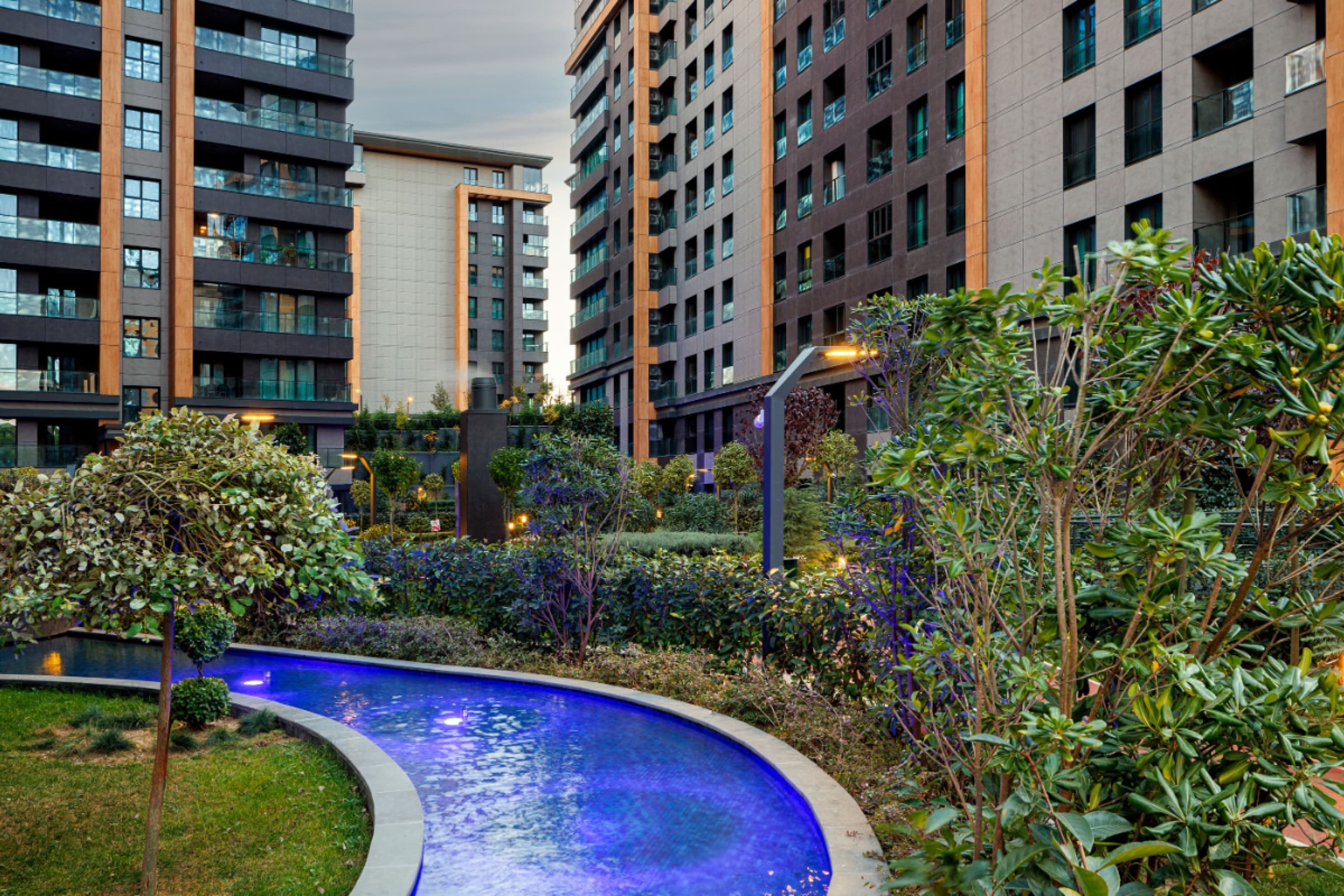 Illuminated garden pool at dusk between two residential blocks, planting grown in