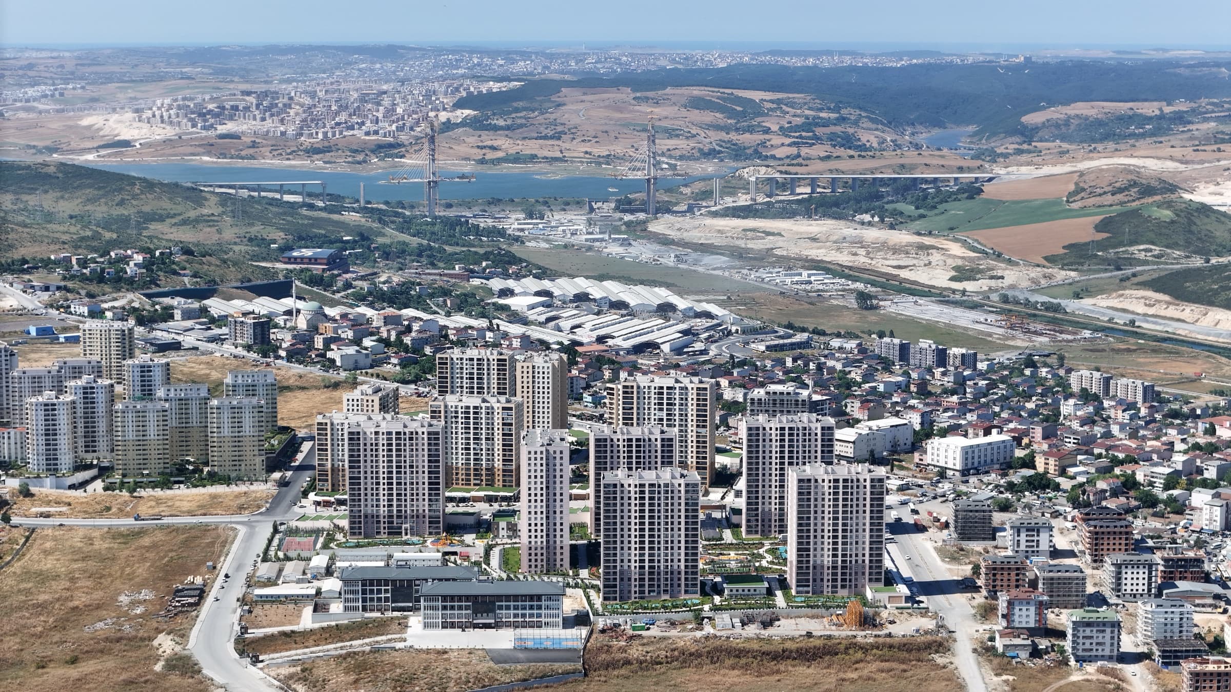 Bizim Evler 11 من الجو نحو الغرب: الأبنية وأرضها المنسّقة تجتمع في حيّ من الشوارع المنخفضة، ومياه مفتوحة وأعمدة جسر عند الأفق