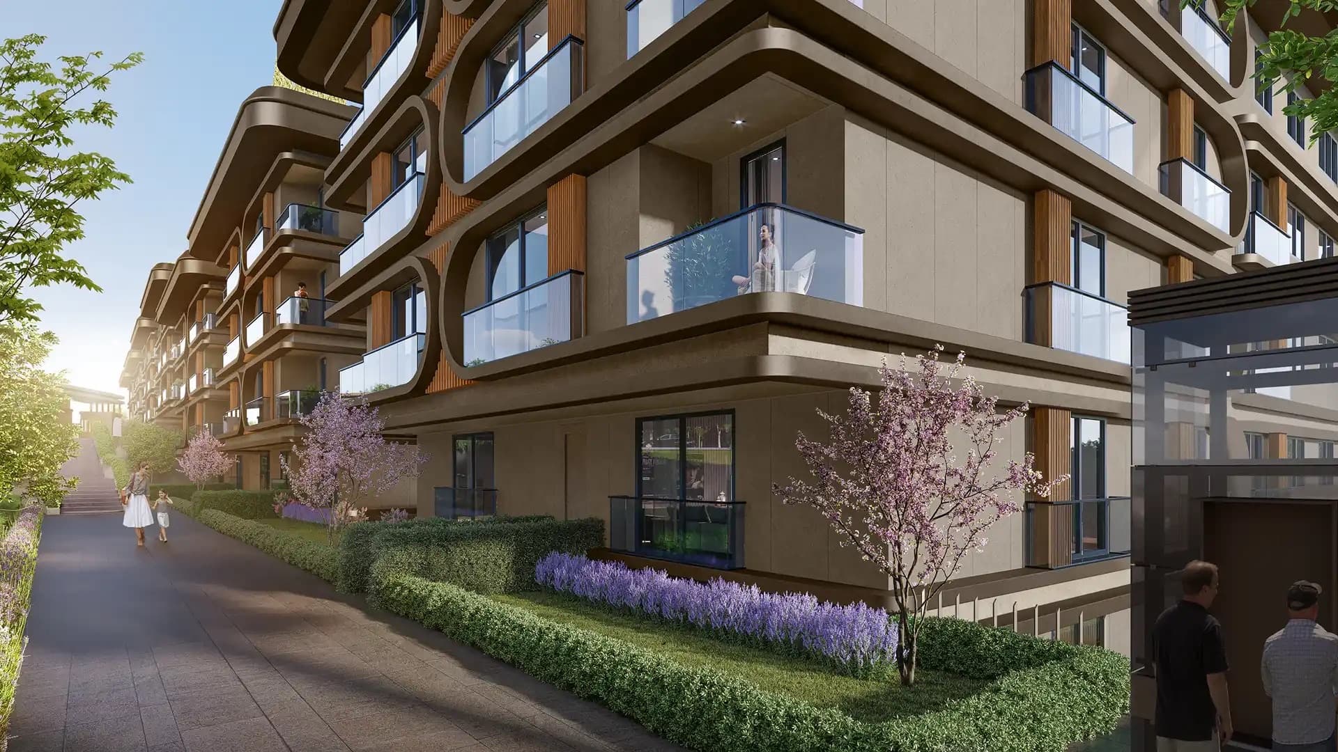The approach to Boğaz Çamlıca at street level, past clipped hedging, lavender and blossom trees, the block's oval balcony frames rising above the footpath