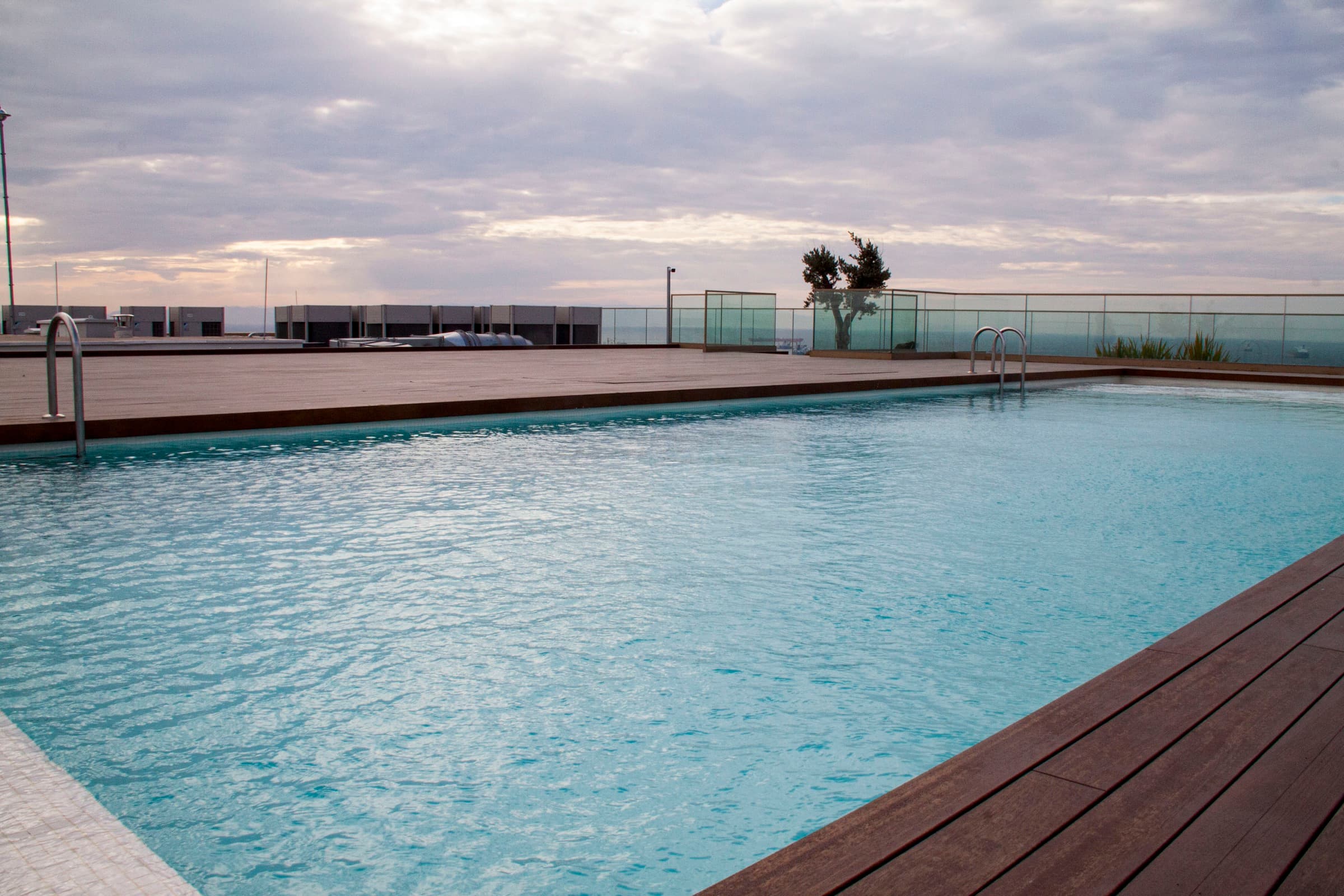 Rooftop swimming pool with timber decking and a glass balustrade, the sea visible under an overcast sky