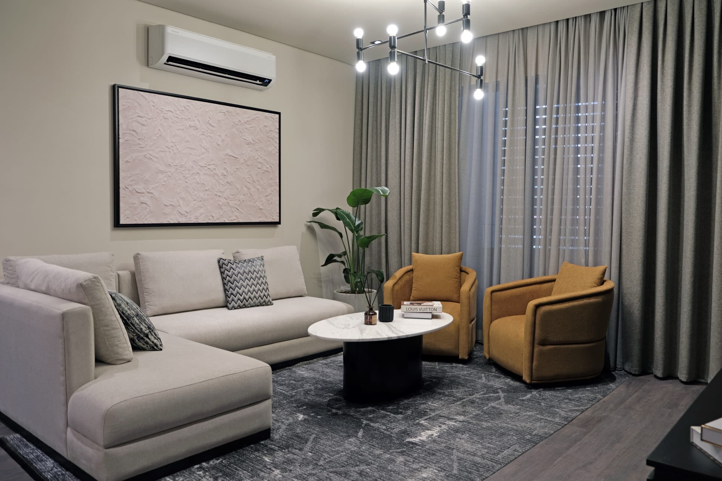 Living room in the furnished show apartment at Dia Centro, with a pale corner sofa, ochre armchairs and a round marble table against full-height curtains
