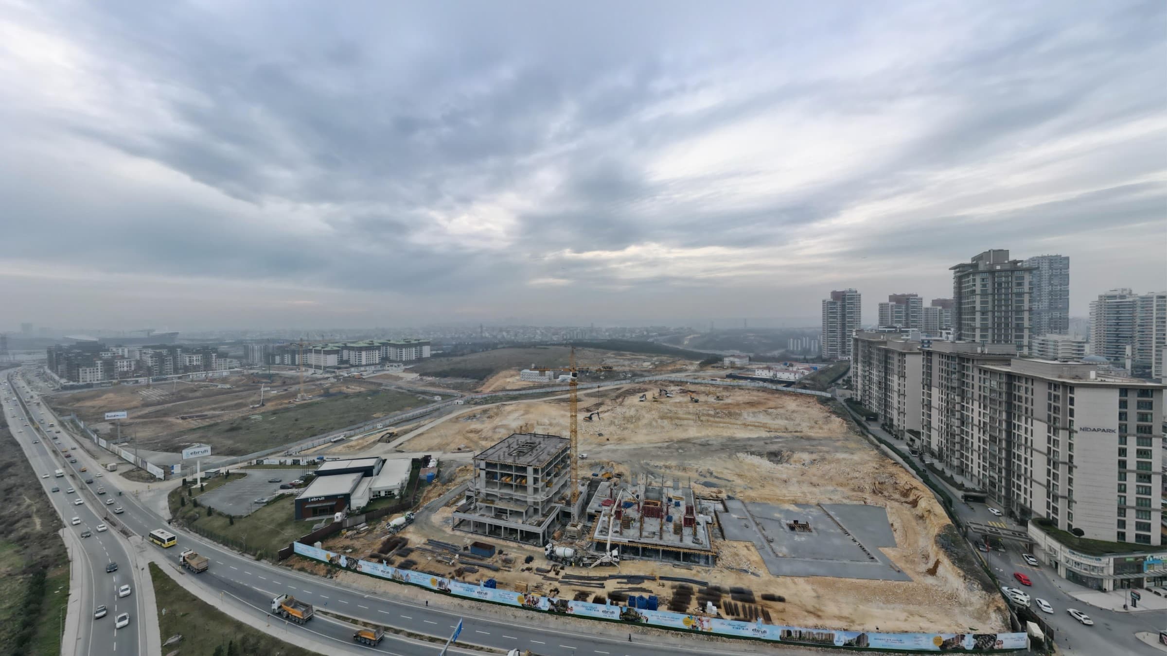 Drone photograph of the Ebruli Başakşehir site in January 2025 under a heavy grey sky: foundations and the first concrete frames rising on bare ground beside the motorway, a tower crane over them, and the existing towers of Başakşehir along the right-hand skyline