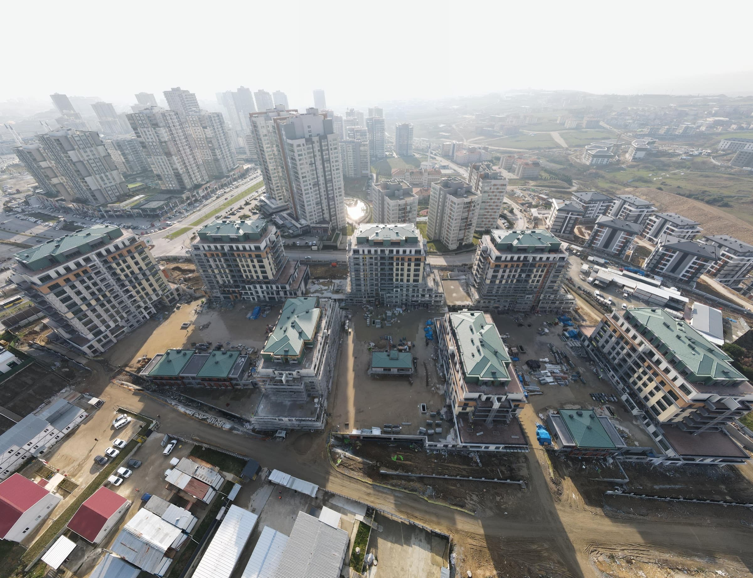 Drone photograph of Ebruli Kayaşehir in January 2025: the blocks structurally complete and clad, their green pitched roofs in place, standing on ground still being worked, with the towers of Kayaşehir receding into haze behind