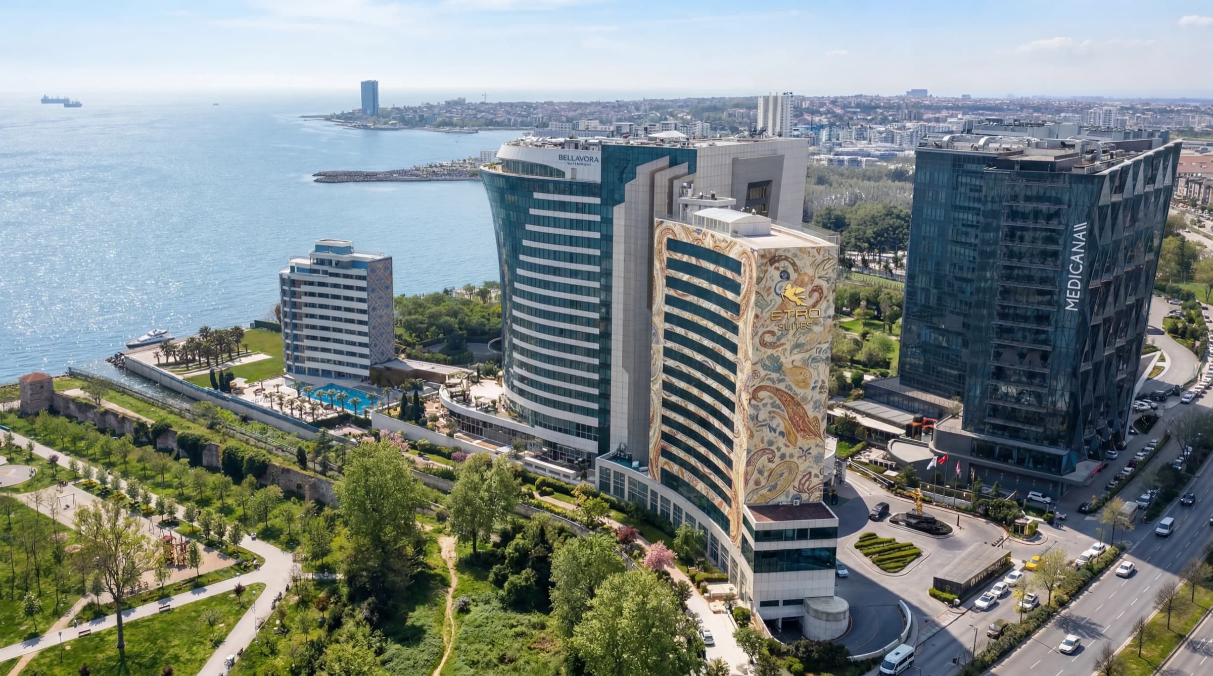 منظر جوي نهاري لمجمّع Bellavora في Ataköy: برج ETRO Suites المكسو بنقش الـ paisley حاملاً شعاره الذهبي، وبرج Bellavora Waterfront الزجاجي يلتفت نحو البحر، والطريق الساحلي والحديقة الساحلية أمامهما، وبحر مرمرة يمتد إلى الأفق