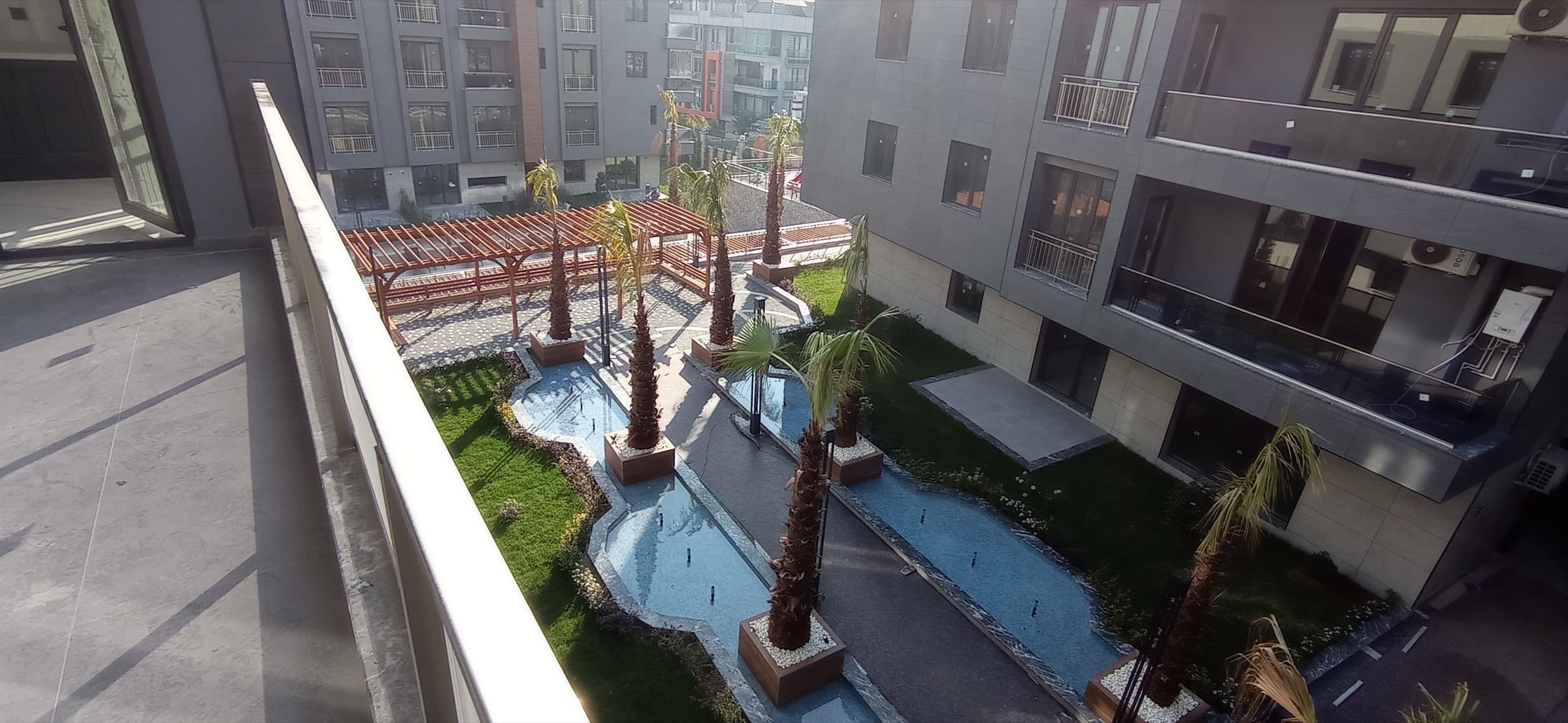View down from a private balcony over the landscaped inner courtyard, with a shallow blue water channel, palm trees in timber planters, lawn and a red pergola between the grey-clad blocks
