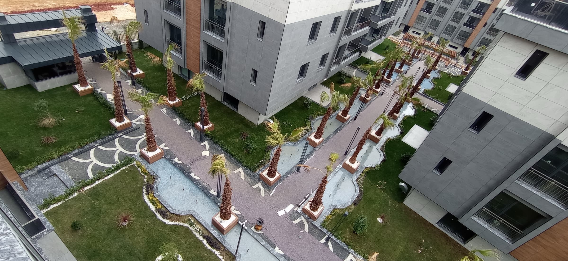 High view over the development's landscaped gardens, showing curving stone-edged water channels, lawns, patterned paving and a double avenue of palms running between the residential blocks