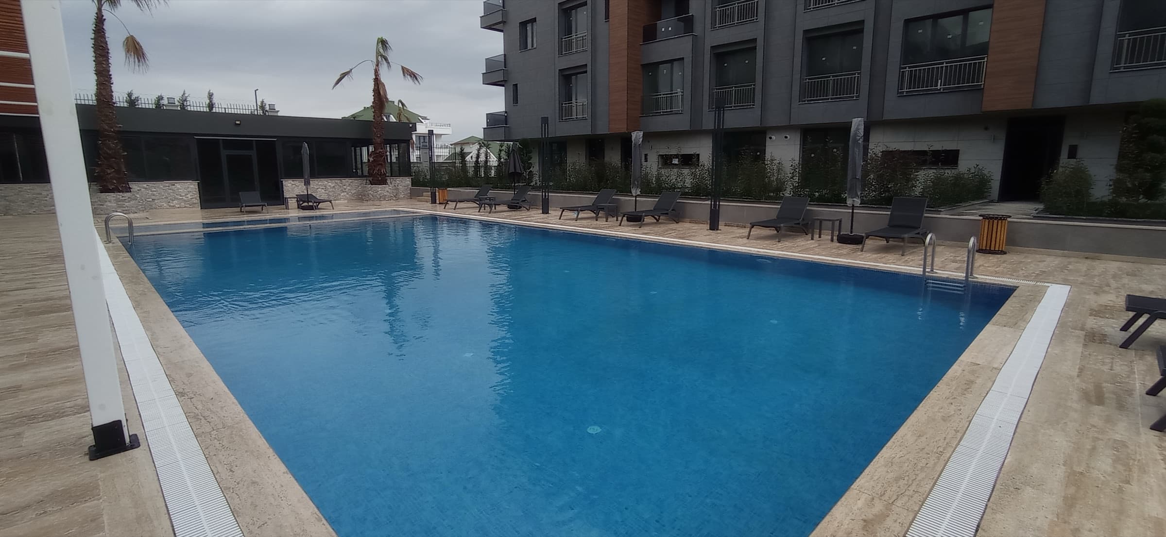 The outdoor swimming pool at Marin City seen the length of its travertine deck, grey sun loungers and folded parasols along the far edge, the single-storey social building to the left and a residential block with planted balconies behind