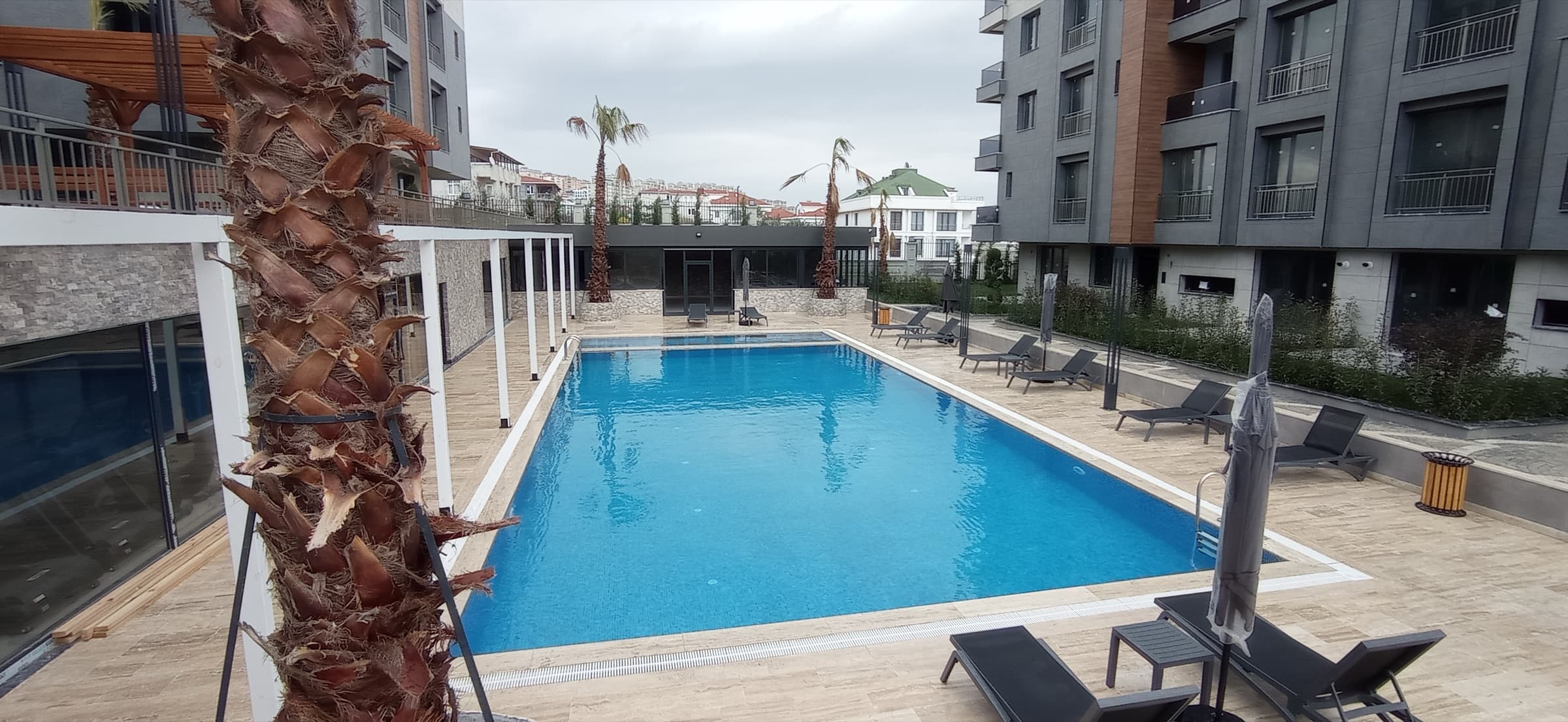 The outdoor swimming pool filled and in use, with travertine decking, grey sunloungers and closed parasols, a white pergola to the left and the residential block rising behind