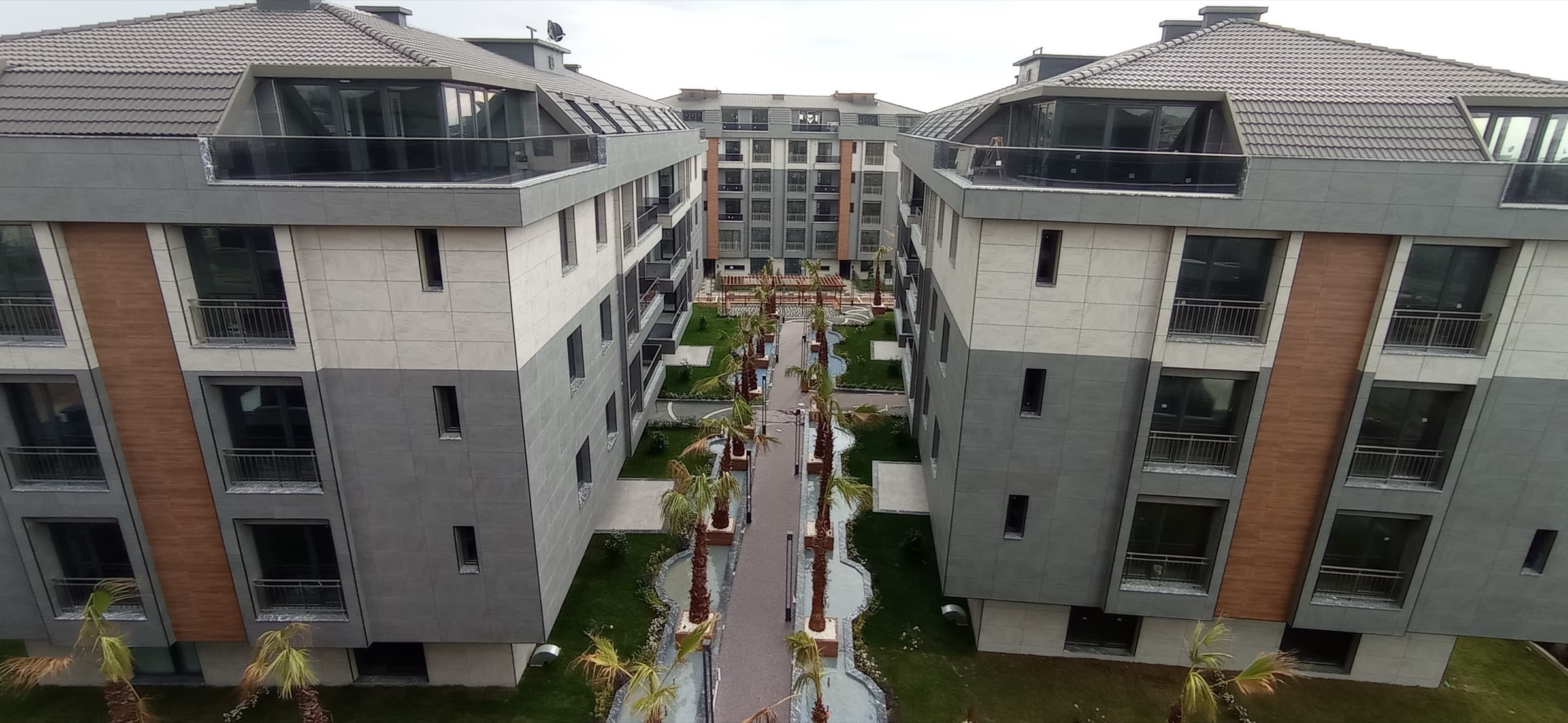 Elevated view between two low-rise blocks with grey stone and timber-effect cladding and glazed rooftop penthouses, looking along the central paved walkway lined with palms towards a taller block behind