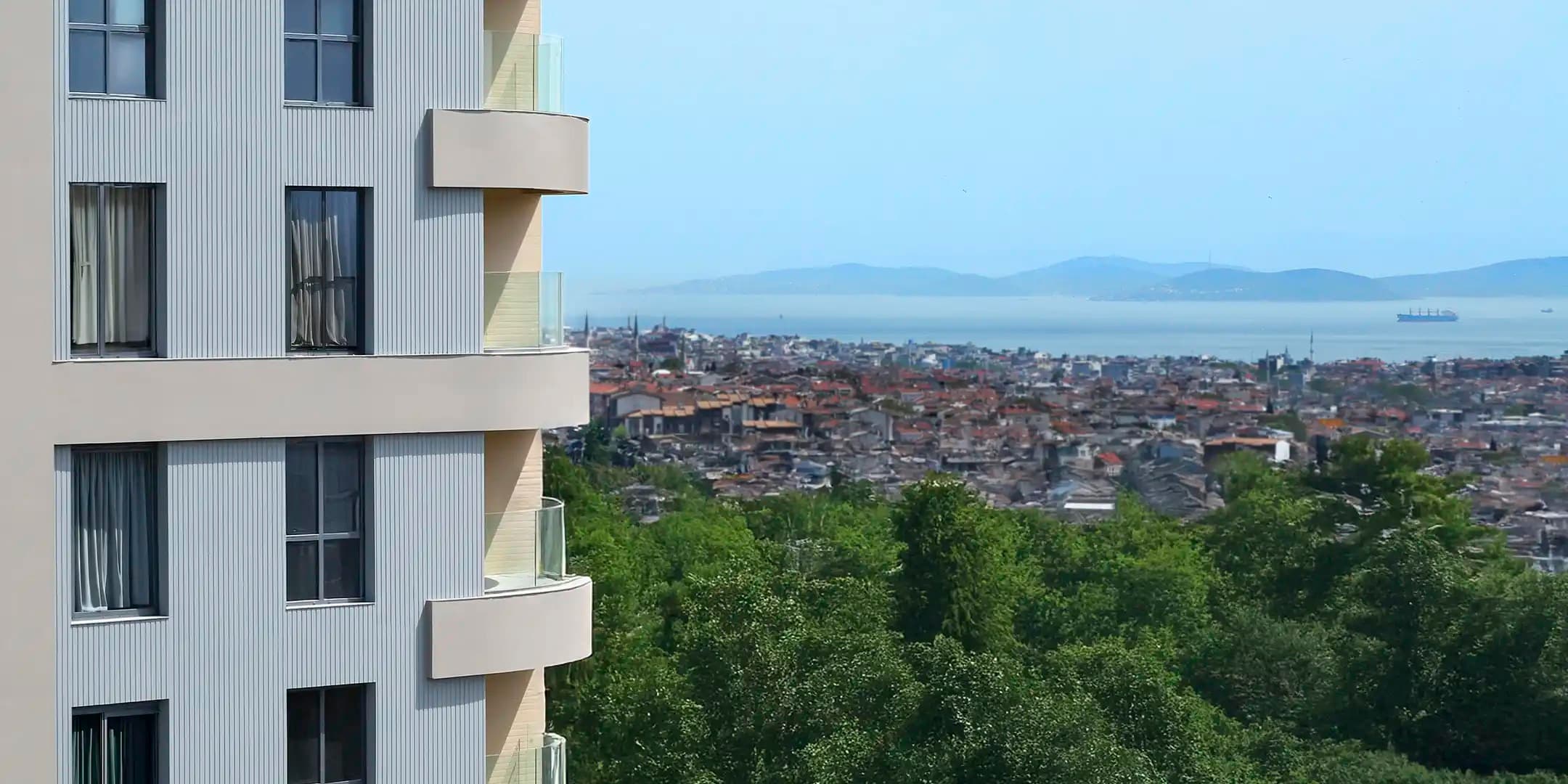 RAMS City Haliç'in üst katlarından manzara: sol kenarda binanın kendi kavisli balkonları, suya doğru alçalan İstanbul çatıları, demirlemiş bir yük gemisi ve karşı kıyıdaki tepeler