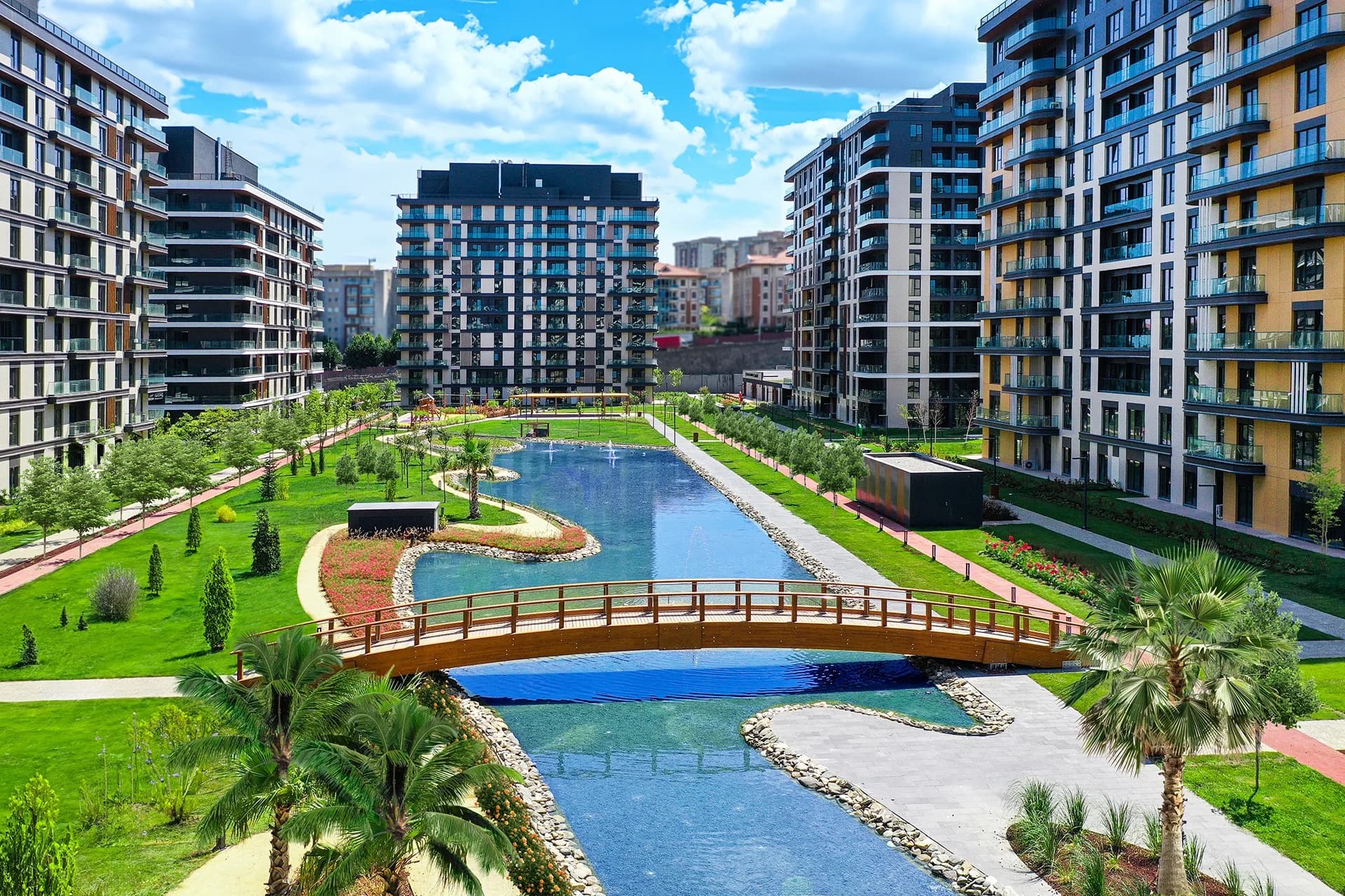 RAMS Garden Bahçelievler'de 210 m uzunluğundaki biyolojik göletin tamamına yüksekten bakış: önde göleti geçen ahşap yaya köprüsü, iki yanda uzanan çim alanlar, palmiyeler ve budanmış bitkilendirme, iki kıyıyı da kuşatan teslim edilmiş daire blokları