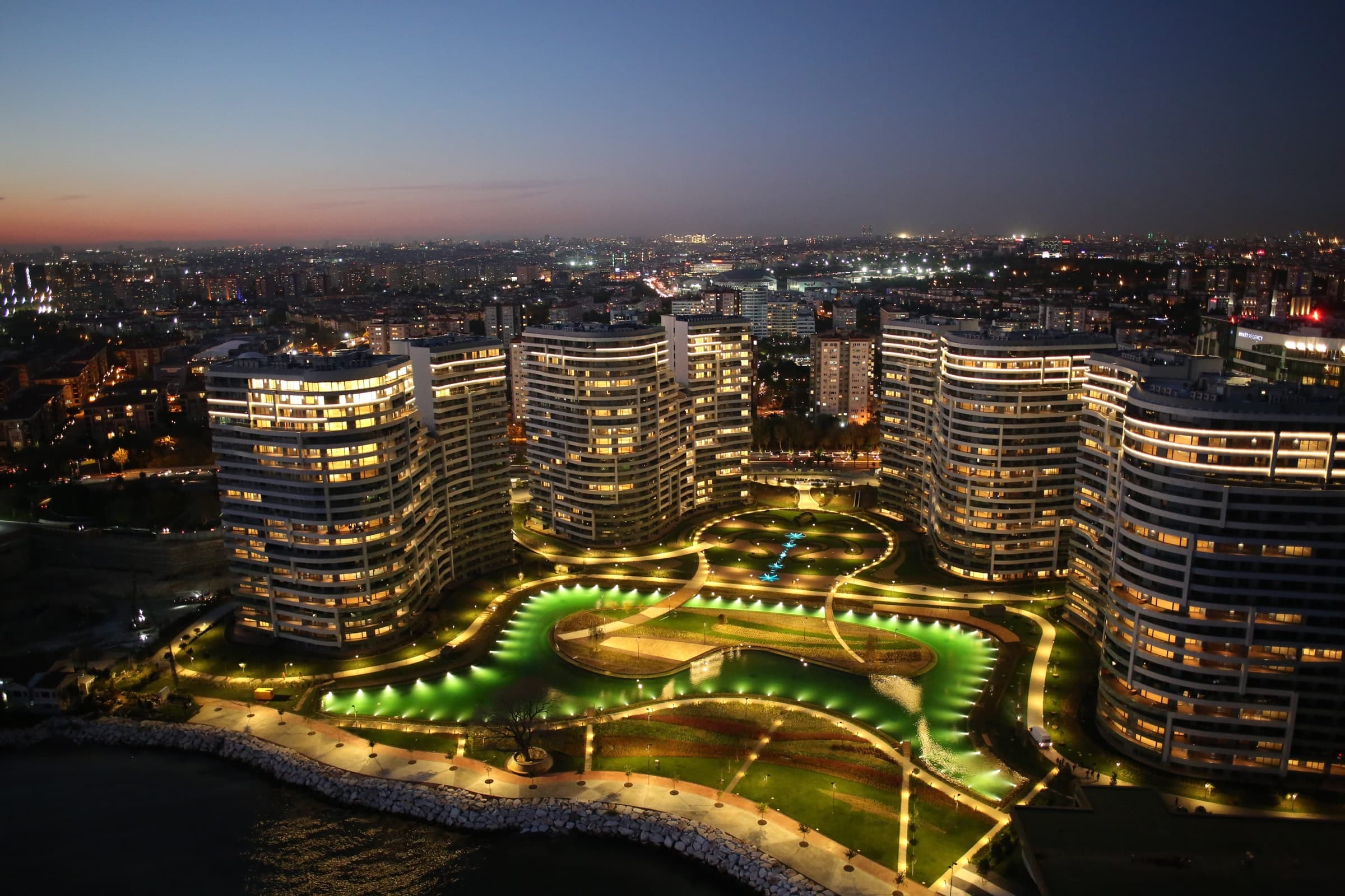 SeaPearl Ataköy from the air at blue hour: the curved blocks set back behind their own rock shoreline on the Marmara, the illuminated gardens and water features between them, and the lights of Istanbul beyond