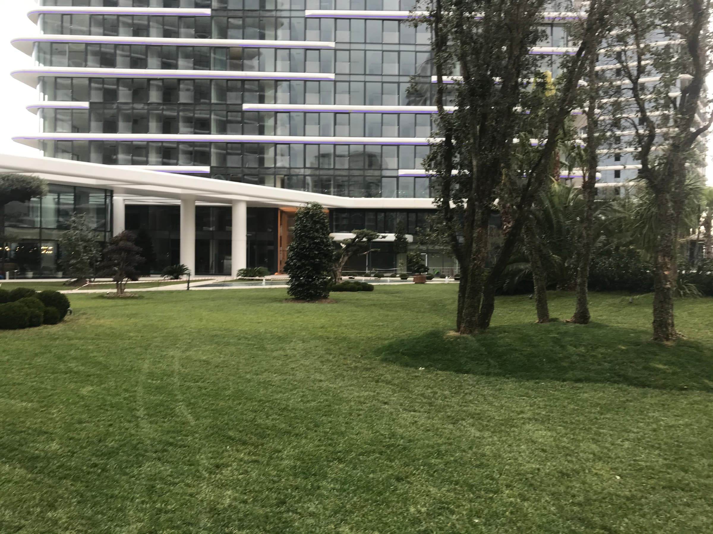 The entrance to Yedi Mavi: a white canopy on slender columns over a timber-lined doorway in the glazed podium, approached across the lawn beneath the residences above