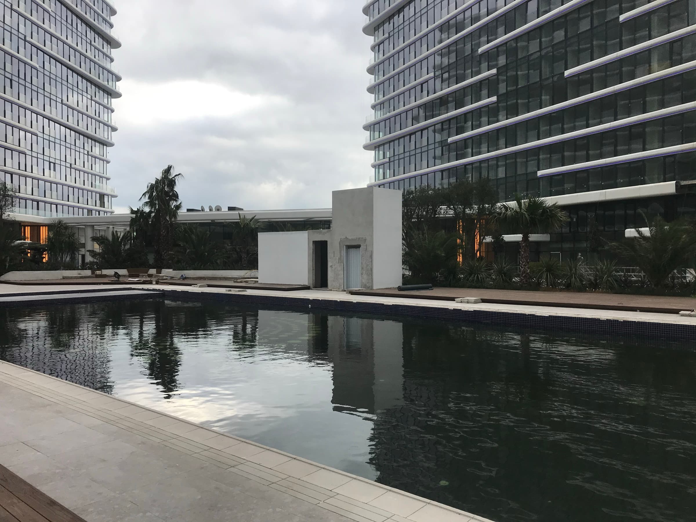 The outdoor pool at Yedi Mavi between two residential blocks, its dark tiled edge holding the reflection of the façades, with palms and timber decking along the far side