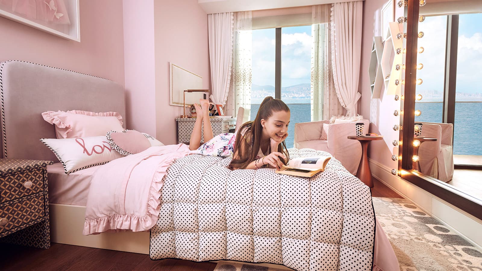 Pink-themed child's bedroom with a girl reading on the bed beside a window overlooking the sea